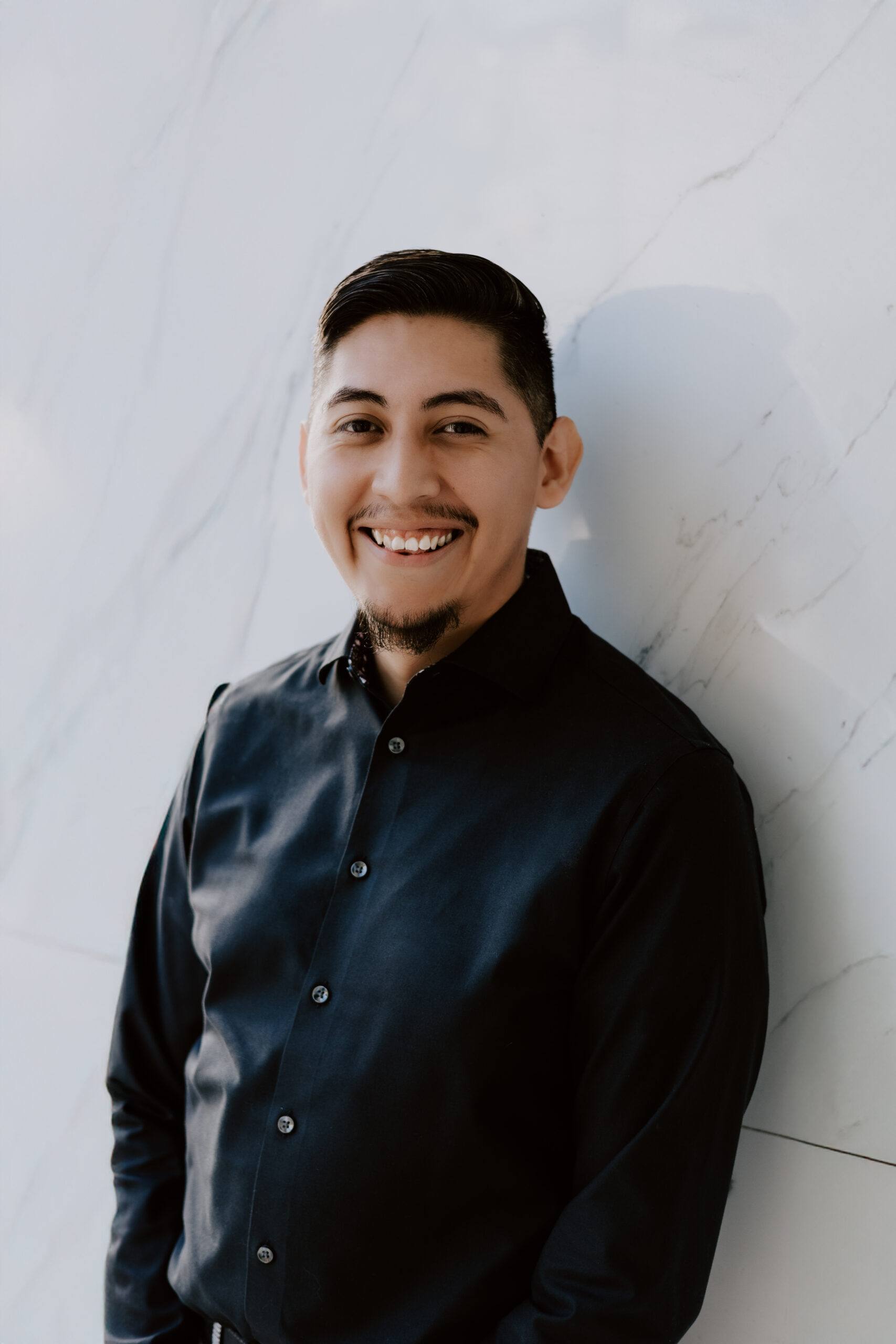 headshot of bilingual male voiceover artist Miguel Anaya standing in front of a white background wall wearing a black button down shirt and smiling at the camera