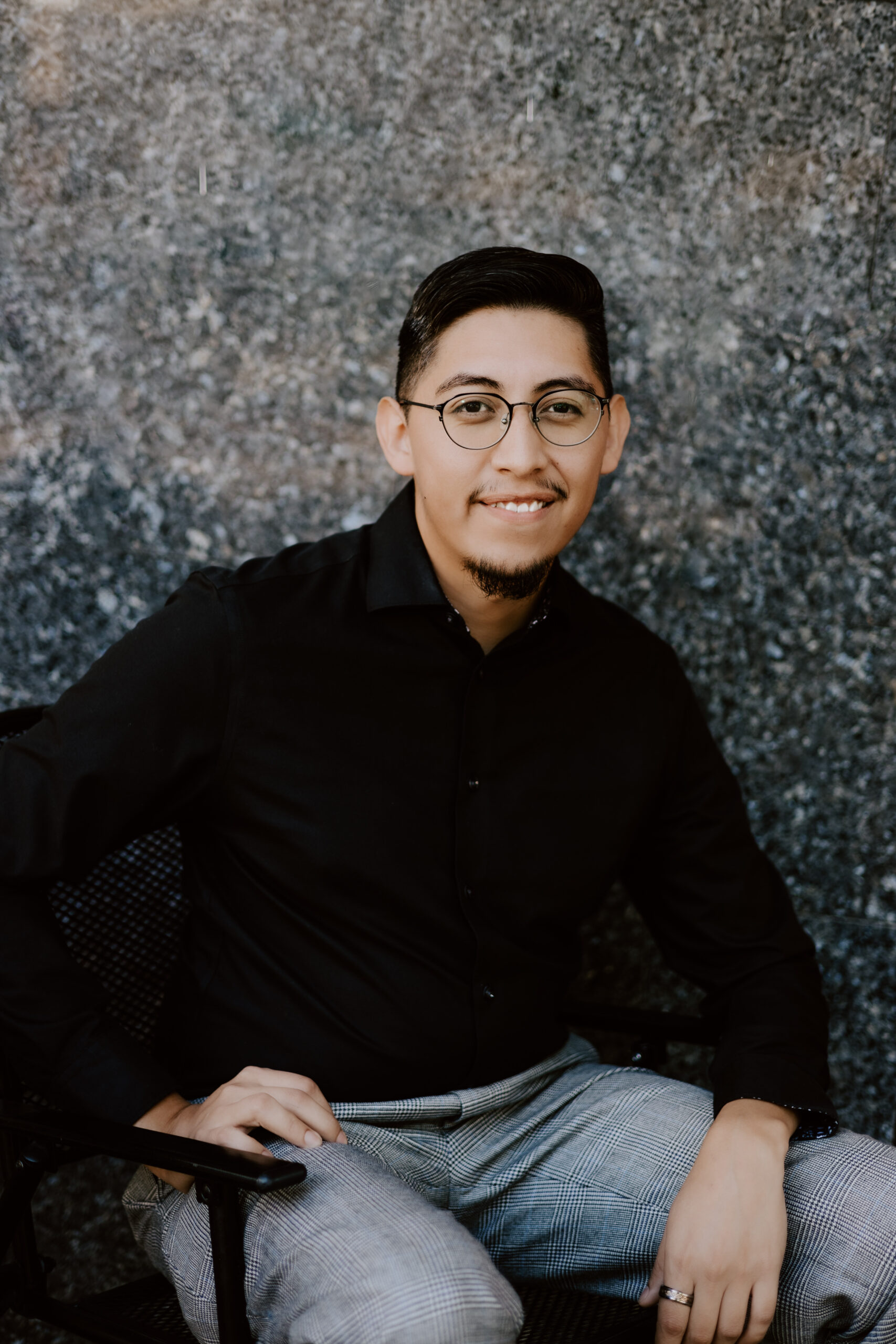 retrato de Miguel Anaya, locutor masculino bilingüe, de pie frente a una pared de fondo gris y blanco, con camisa negra de botones, gafas y sonriendo a la cámara.