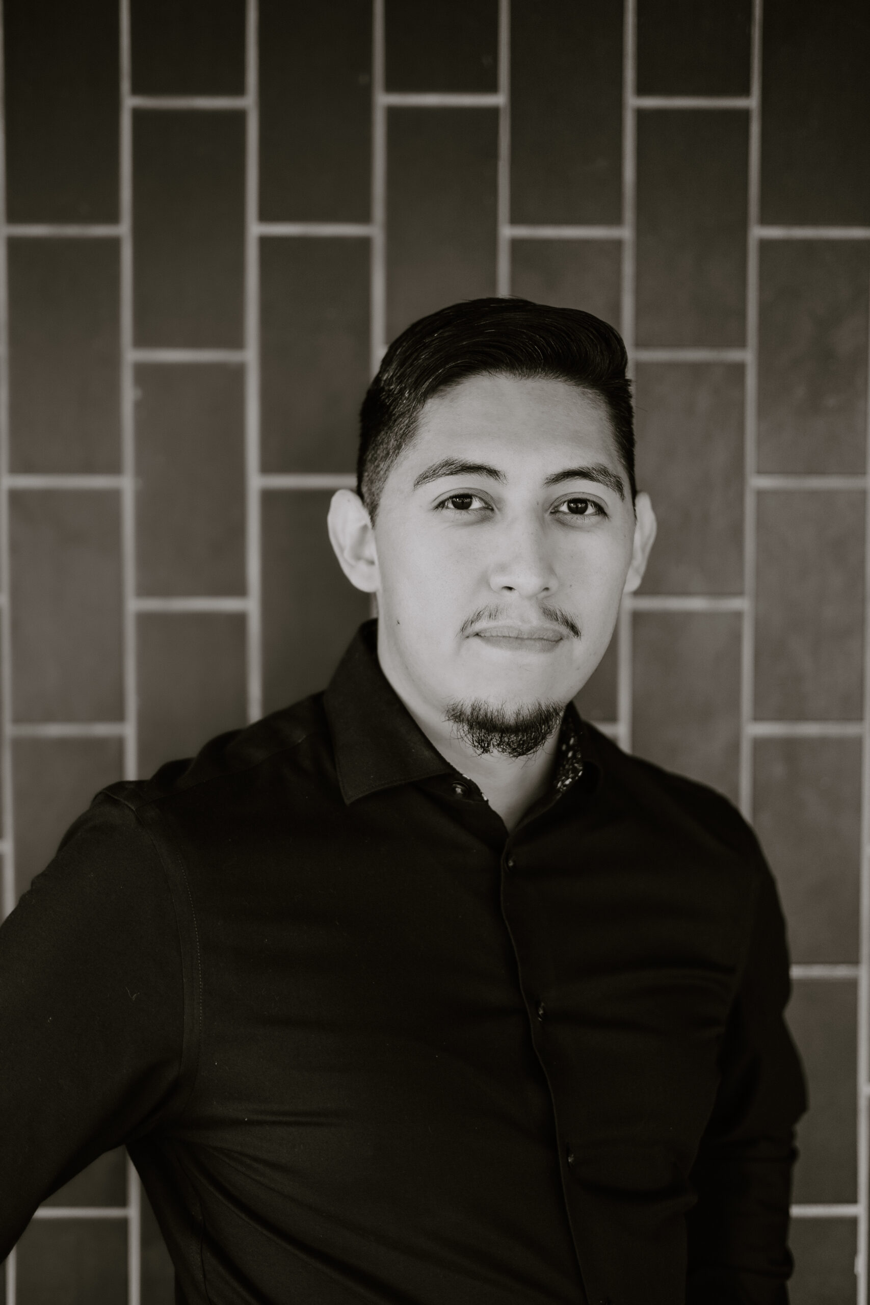 black and white headshot of bilingual male voiceover artist Miguel Anaya standing in front of a blue tile wall wearing a black button down shirt